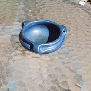 May include: A small, round, blue ceramic bowl with a glossy finish. The bowl has a wide opening and two small handles on opposite sides. The interior is a lighter shade of blue, and the exterior is a darker, more saturated blue.