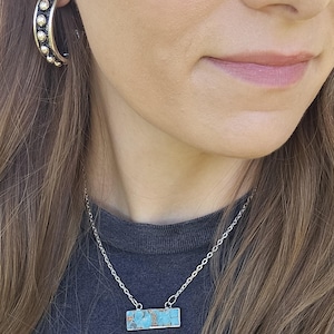 May include: A silver necklace with a rectangular turquoise pendant. The necklace is worn with a dark gray t-shirt featuring a large white and yellow daisy graphic. Silver hoop earrings are also visible.