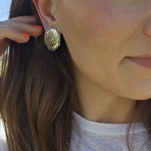 May include: A silver-toned stud earring with a geometric design is visible. The earring has an octagonal shape with a detailed pattern in the center. The person is wearing a white t-shirt and a beaded necklace.