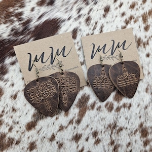 May include: A pair of brown leather earrings shaped like guitar picks with a geometric pattern. The earrings are on a white background with the text "www.theleathermarket.com".