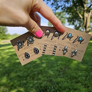 May include: A selection of silver-toned stud earrings on display. The earrings feature various designs, including floral, teardrop, and geometric shapes. Some studs have turquoise accents. The earrings are presented on small, brown cards.
