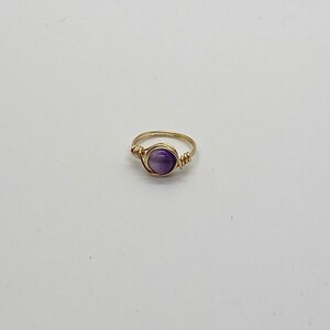 Könnte beinhalten: Ein goldfarbener Ring mit einem violetten Amethyst-Edelstein, der mit Draht umwickelt ist.
