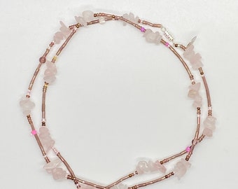 Puede incluir: Un collar de doble cadena hecho con trozos de cuarzo rosa y pequeñas cuentas marrones. El collar tiene un cierre plateado.