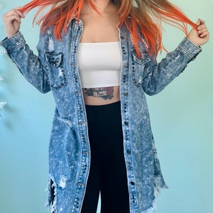 May include: A light blue denim jacket with distressed details and black buttons. The jacket has a relaxed fit and is slightly oversized. The model is wearing a white tank top and black pants underneath the jacket.
