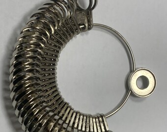 Könnte beinhalten: Ein silberner Ringgrößenmesser mit einer Reihe von abgestuften Ringen, der zum Messen der Fingergrößen verwendet wird. Der Größenmesser ist aus Metall und hat eine kreisförmige Form.