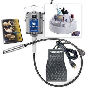 May include: A gray Foredom SR rotary tool with a foot pedal, a black cord, and a gray stand with a variety of bits and a tube of lubricant. The tool is labeled "Foredom SR" and has a speed control dial. The stand has a circular base with a tiered design and a central cylinder. The bits are arranged in rows around the cylinder. The tube of lubricant is labeled "Foredom Lubricant".