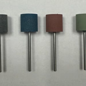 Puede incluir: Cuatro tambores de lijado con superficies abrasivas de diferentes colores montados en ejes metálicos. Los tambores son grises, azules, marrones y verdes.