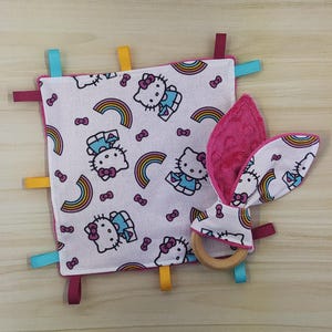 May include: A baby comfort set featuring a white square blanket and a bunny ear teether. The blanket has a print of a cartoon cat, rainbows, and bows. The teether has a wooden ring and pink plush ears. Colorful ribbons are attached to the blanket.