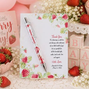 May include: A white thank you card with a strawberry and green leaf border, featuring the text Thank You, For showering us with love, With Love, Amanda & Kevin, May 18, 2025, paired with a clear pen containing floating red strawberry charms.