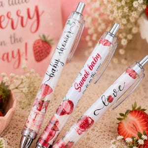 May include: Three retractable ballpoint pens with white barrels featuring strawberry illustrations and text. The pens display the phrases baby shower, Sweet baby is on the way, and love in black and red script fonts, with silver metal clips and tips.