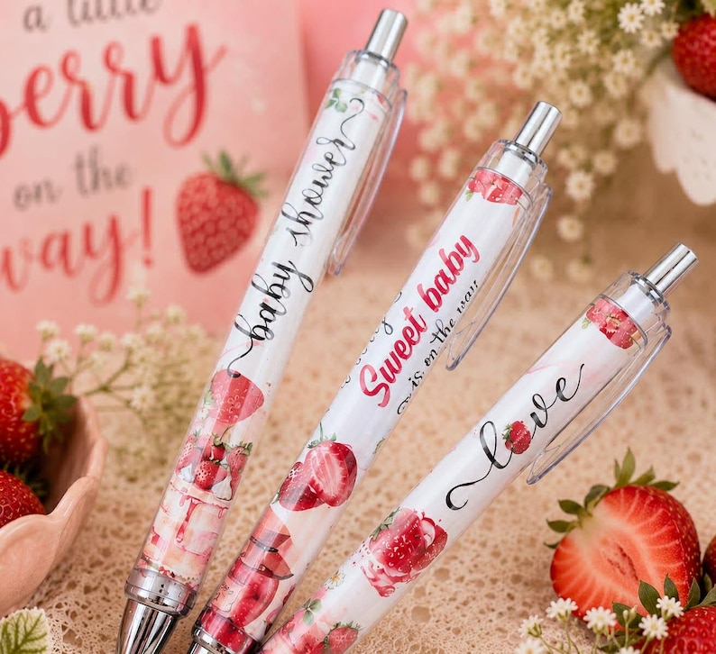 May include: Three retractable ballpoint pens with white barrels featuring strawberry illustrations and text. The pens display the phrases baby shower, Sweet baby is on the way, and love in black and red script fonts, with silver metal clips and tips.