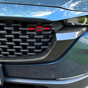 May include: A dark teal car front grille featuring a black honeycomb pattern with three horizontal red accent bars positioned on the upper left side. The grille is framed by a glossy black trim piece that extends toward the headlight assembly.