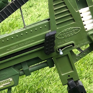 May include: Green plastic toy rifle with a black grip and a silver bolt. The rifle has the words "TOPPERS" and "RIFLE BOLT" printed on it. The rifle is on a green grassy surface.