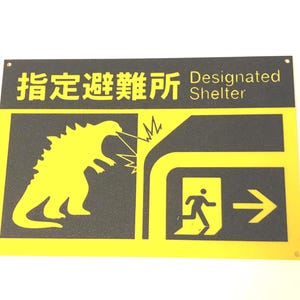 Puede incluir: Un letrero amarillo y negro con las palabras "Designated Shelter" y caracteres japoneses. El letrero presenta una silueta amarilla de un monstruo que respira fuego, una persona corriendo hacia una salida y una flecha.
