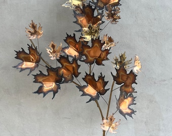 May include: A wall sculpture made of metal leaves in shades of gold, copper, and brown. The leaves are arranged in a cascading pattern on a thin metal stem.