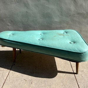 May include: A turquoise blue, triangular shaped, upholstered ottoman with button tufting and wooden legs.