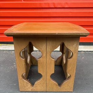 May include: A wooden side table with a square top and decorative cutouts on the sides. The table is made of light brown wood and has a simple, yet elegant design. The table is suitable for a living room or bedroom.