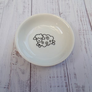 Pode incluir: Um prato de cerâmica branco com um contorno preto de uma ovelha com um rosto sorridente. A ovelha tem lã encaracolada.