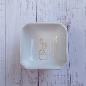 Könnte beinhalten: Eine weiße Keramik Schale mit einem goldenen Kaktus-Design. Die Schale ist quadratisch mit abgerundeten Ecken und hat einen leicht erhöhten Rand.