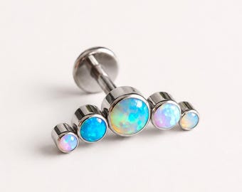 May include: A silver-toned metal labret stud featuring a curved, fan-shaped arrangement of five round, iridescent opals. The stones graduate in size from the centre outward, displaying flashes of blue, pink, and white light against a metallic setting.