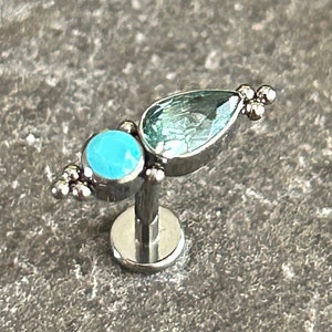 May include: A silver stud earring with a turquoise round stone and a light blue, teardrop-shaped stone. The earring has a flat base and a post for piercing. Small silver beads decorate the design.