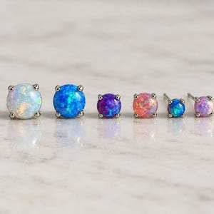 May include: A set of six round stud earrings featuring iridescent opal-like stones in various colors including white, blue, purple, and pink. Each stone is set in a silver-toned metal prong setting, with two earrings showing the attached metal posts.
