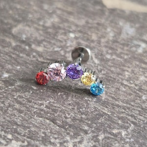 May include: A curved metal body jewellery stud featuring five round faceted gemstones in a row. The stones are arranged in a gradient of colours including red, light pink, purple, yellow, and blue, set into a silver-toned metal base with a flat back.