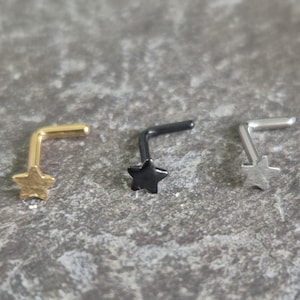 Puede incluir: Tres piercings nasales con forma de estrella, en oro, negro y plata. Cada uno tiene un pequeño adorno de estrella y un poste en forma de L. Los piercings se muestran sobre una superficie gris texturizada.