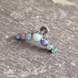 May include: A curved barbell body jewellery piece featuring seven round, iridescent opal stones set in a silver-toned metal frame. The stones graduate in size, with the largest opal positioned in the centre, flanked by smaller opals that display blue, pink, and white flashes of colour.