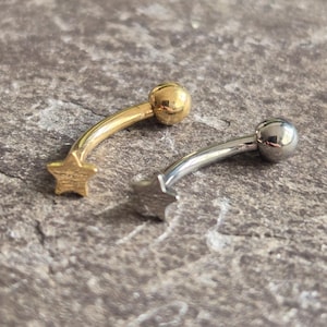 May include: Two curved eyebrow rings. One is gold-colored with a star charm and a ball end. The other is silver-colored with a ball end and a star charm. Both are on a textured gray surface.