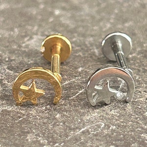 Puede incluir: Dos pendientes de media luna y estrella, uno dorado y otro plateado, sobre una superficie gris texturizada.