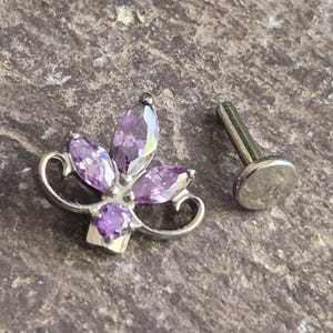 May include: A silver-toned body jewellery piece featuring a decorative top with four marquise-cut purple stones and one round purple stone arranged in a floral pattern, accompanied by a separate flat-back post with a circular base for secure attachment.