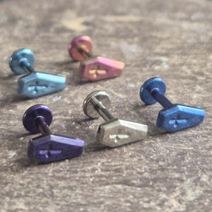 May include: A collection of coffin-shaped stud earrings in various colors, including blue, purple, pink, and silver. Each earring features a cross design on the coffin. The earrings are displayed on a textured surface.