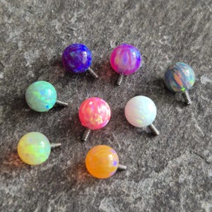 Puede incluir: Colección de siete pendientes de bola con efecto ópalo en varios colores, incluyendo azul, rosa, verde y naranja. Cada pendiente tiene un poste de rosca plateado. Los pendientes están dispuestos sobre una superficie gris texturizada.