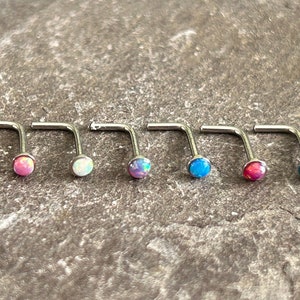 Puede incluir: Seis piercings nasales en forma de L con piedras de ópalo de colores. Los piercings están dispuestos en fila, mostrando una variedad de colores que incluyen rosa, blanco, morado, azul, rojo y turquesa. Las joyas están sobre una superficie gris texturizada.