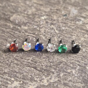 May include: Six small stud earrings with various coloured gemstones. The gemstones are red, iridescent, blue, white, green, and black. Each earring has a silver post. They are displayed on a textured grey surface.