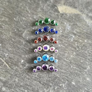 May include: Six curved body jewellery pieces arranged vertically, each featuring five bezel-set round gemstones in varying colours including green, blue, red, white, light blue, and purple, all mounted on a polished silver-tone metal base.