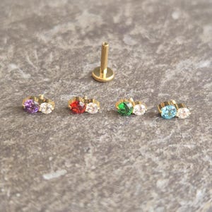 May include: Gold-colored body jewelry with a flat back and a post. Four pieces feature a gold setting with two gemstones in various colors: purple, red, green, and blue. A single post is also visible.