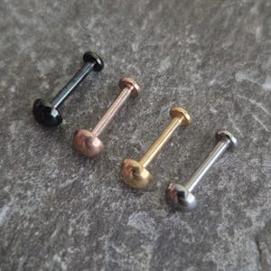 Puede incluir: Cuatro piercings tipo labret con base plana y extremo redondeado. El conjunto incluye uno negro, uno de oro rosa, uno de oro amarillo y uno de tono plateado. Cada pieza presenta un poste recto que conecta la base plana con la parte superior.
