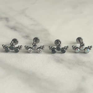 May include: Three black titanium body jewelry pieces with different colored gems. The first piece has green gems, the second has clear gems, and the third has black gems.