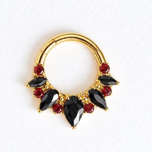 May include: A gold-toned hinged hoop ring featuring a decorative cluster of three pear-shaped black stones and four round red stones arranged in a fan-like pattern along the lower half of the circular metal band.