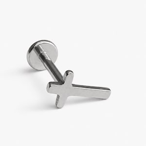May include: A silver-toned metal labret stud body jewelry piece featuring a flat circular back and a cross-shaped front ornament. The cross has smooth, rounded edges and a polished finish, attached to a straight post for secure placement.