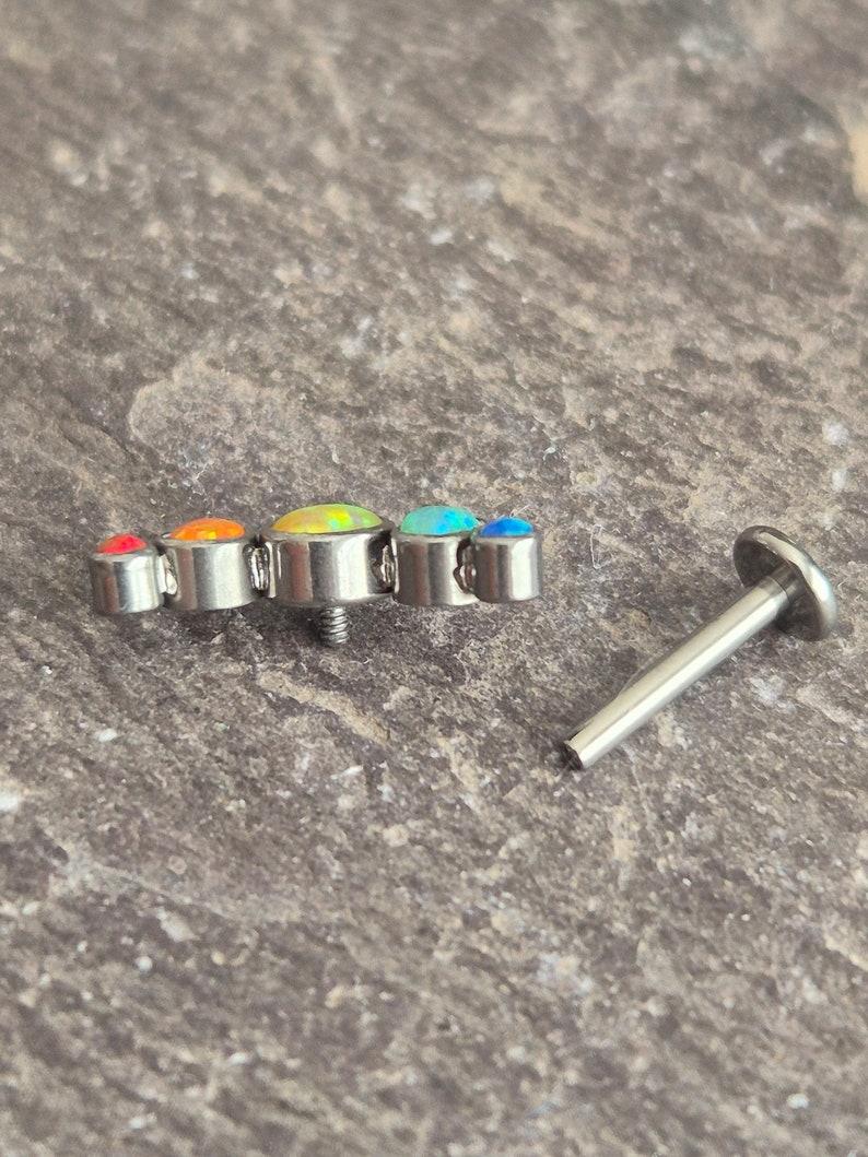 May include: A silver-toned metal body jewellery piece featuring a curved row of five round, multi-coloured cabochon stones in shades of red, orange, yellow, and blue, accompanied by a separate flat-backed metal post with a threaded end.