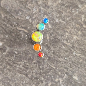 May include: A curved body jewellery piece featuring five round, bezel-set stones in a graduated size arrangement. The stones display vibrant, iridescent colours including blue, light blue, yellow, orange, and red, all set in a polished silver-tone metal.