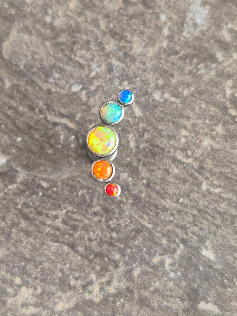 May include: A curved body jewellery piece featuring five round, bezel-set stones in a graduated size arrangement. The stones display vibrant, iridescent colours including blue, light blue, yellow, orange, and red, all set in a polished silver-tone metal.