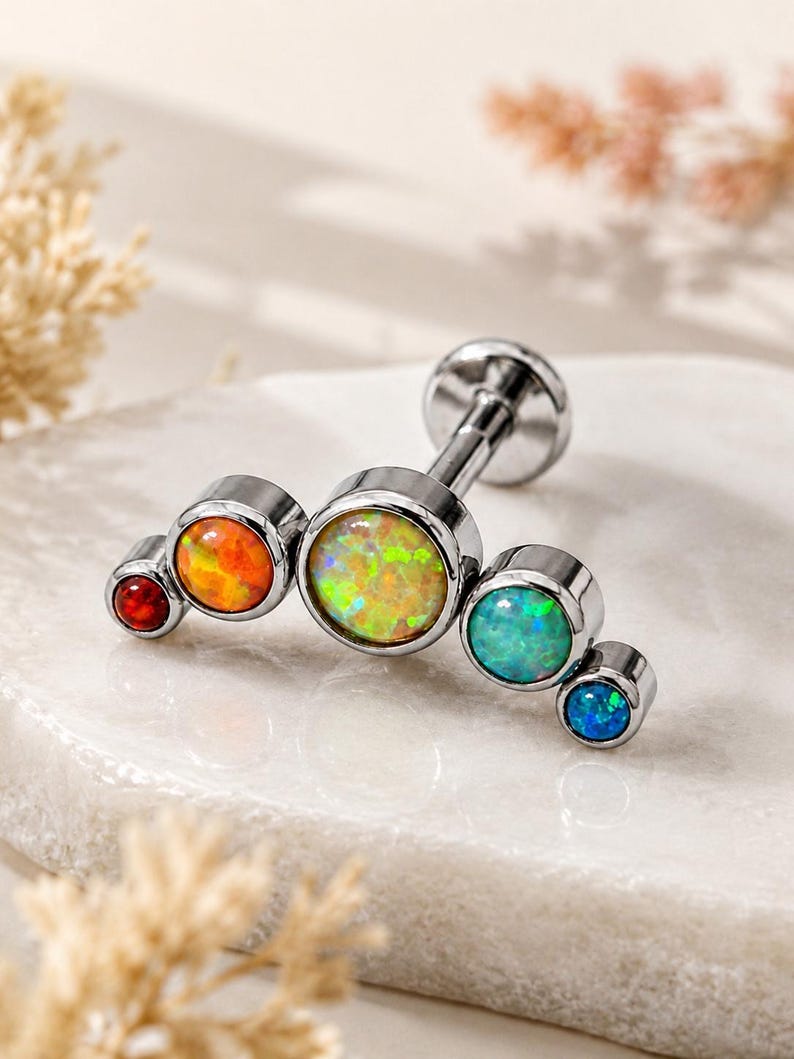 May include: A silver-toned metal labret stud featuring a curved row of five round bezel-set stones in a gradient of colours. The stones transition from a small deep red, to orange, a large central yellow-green, teal, and a small dark blue stone.