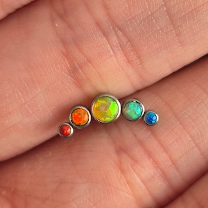 May include: A curved body jewellery piece featuring five bezel-set stones in a gradient of colours. The stones transition from a small red gem, to a larger orange one, a central yellow-green stone, a light green gem, and a small blue stone at the end.