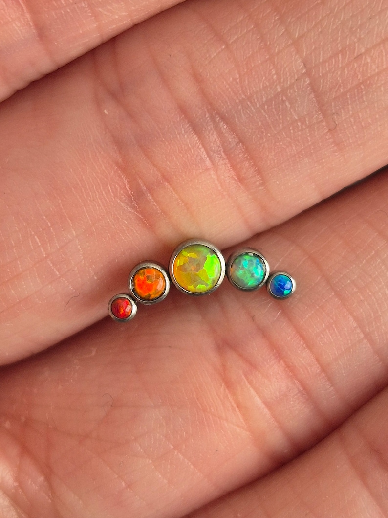 May include: A curved body jewellery piece featuring five bezel-set stones in a gradient of colours. The stones transition from a small red gem, to a larger orange one, a central yellow-green stone, a light green gem, and a small blue stone at the end.
