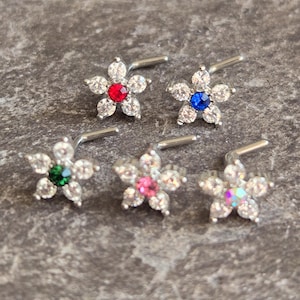 May include: Five flower-shaped nose studs. Each stud features a central gemstone in red, blue, green, pink, and iridescent colors, surrounded by clear crystals. The studs are set on a silver-colored post.