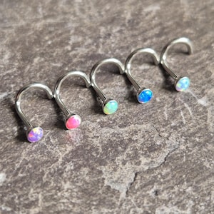 May include: Five nose rings with curved posts and small, round opal stones in various colours: purple, pink, green, blue, and white. The jewellery is made of silver-coloured metal and is displayed on a textured, grey surface.